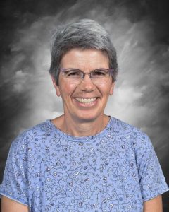 Smiling person with short gray hair, wearing glasses and a floral blue shirt, against a gray background.