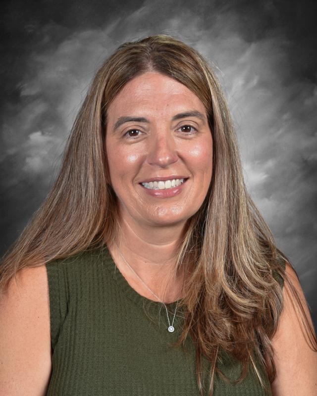 Smiling woman with long brown hair, wearing a green top, against a gray backdrop.
