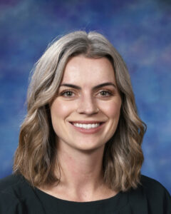 A smiling woman with shoulder-length, wavy gray hair against a blue background. She is wearing a black top.