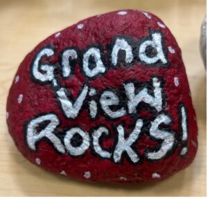 A red-painted rock with the words "Grand View Rocks!" written in white and black, surrounded by white dots.