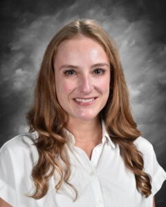 A woman with long, wavy brown hair smiles at the camera, wearing a white shirt against a gray background.