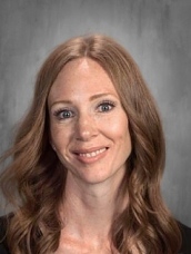 A smiling woman with long, wavy hair and blue eyes poses in front of a gray backdrop. She wears a black top.