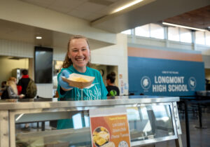 A smiling student in a turquoise shirt serves food at Longmont High School's cafeteria.