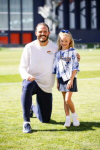 A smiling man in a Broncos shirt kneels beside a girl in a Broncos outfit on a grassy field.
