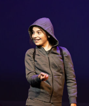 A young girl in a gray hoodie smiles and gestures while performing on stage, with a purple background.