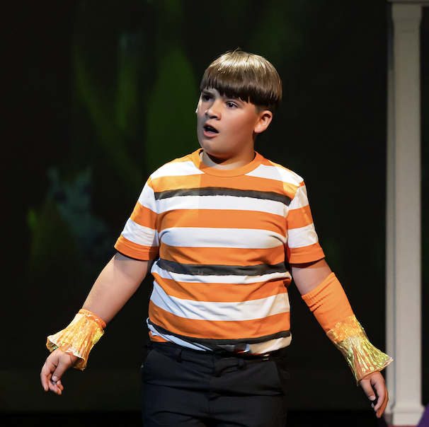 A boy with short hair wears a striped orange and white shirt and yellow sleeves, acting surprised on stage.