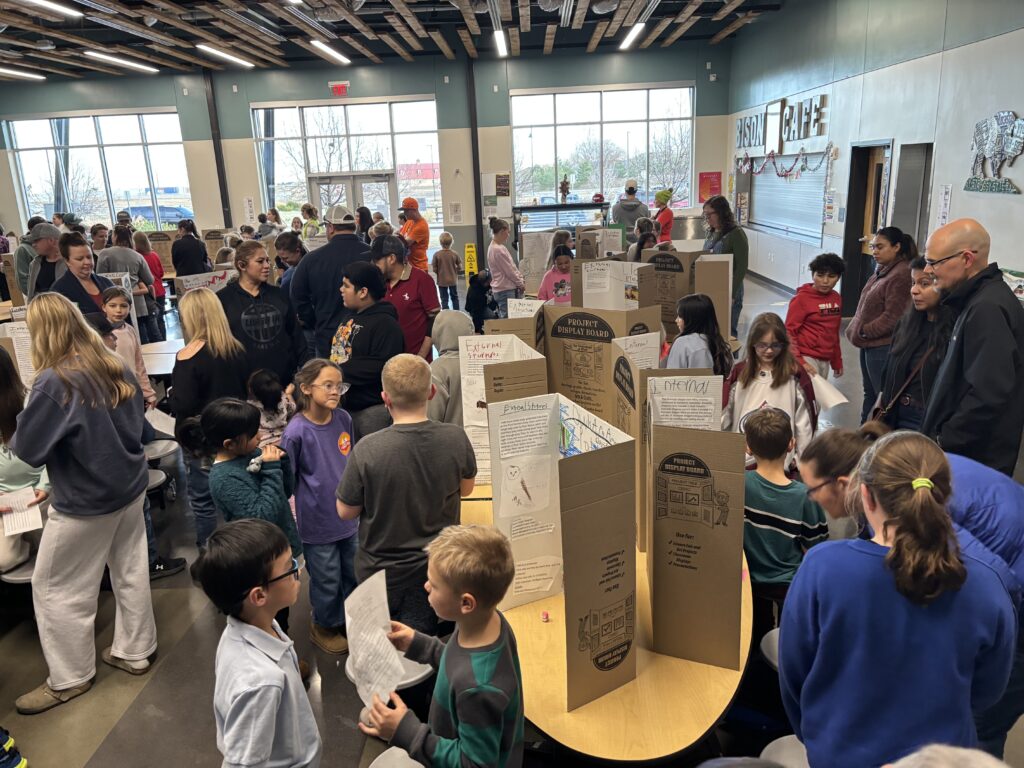 A crowded school event with children and adults interacting around project display boards in a bright, spacious room.