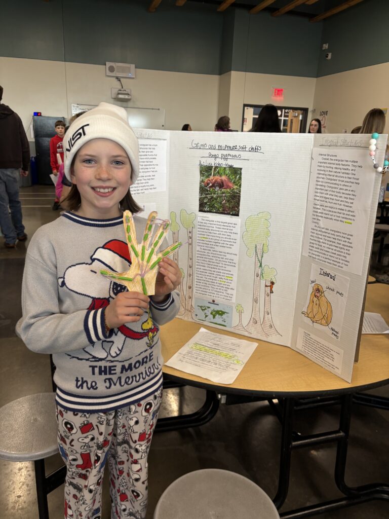 A young girl in a winter hat holds a colorful project about animals, standing in front of a presentation board.