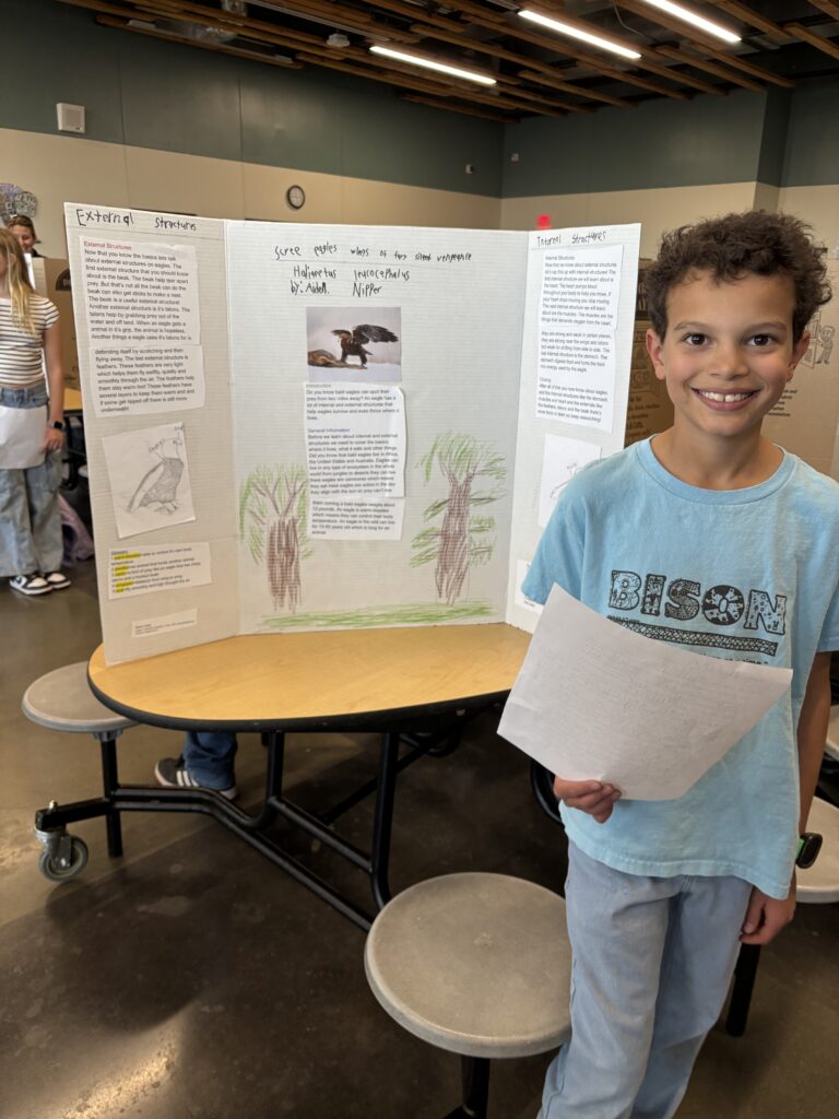 A young boy stands by a science project display board, holding a paper, with illustrations and text about external and internal structures.