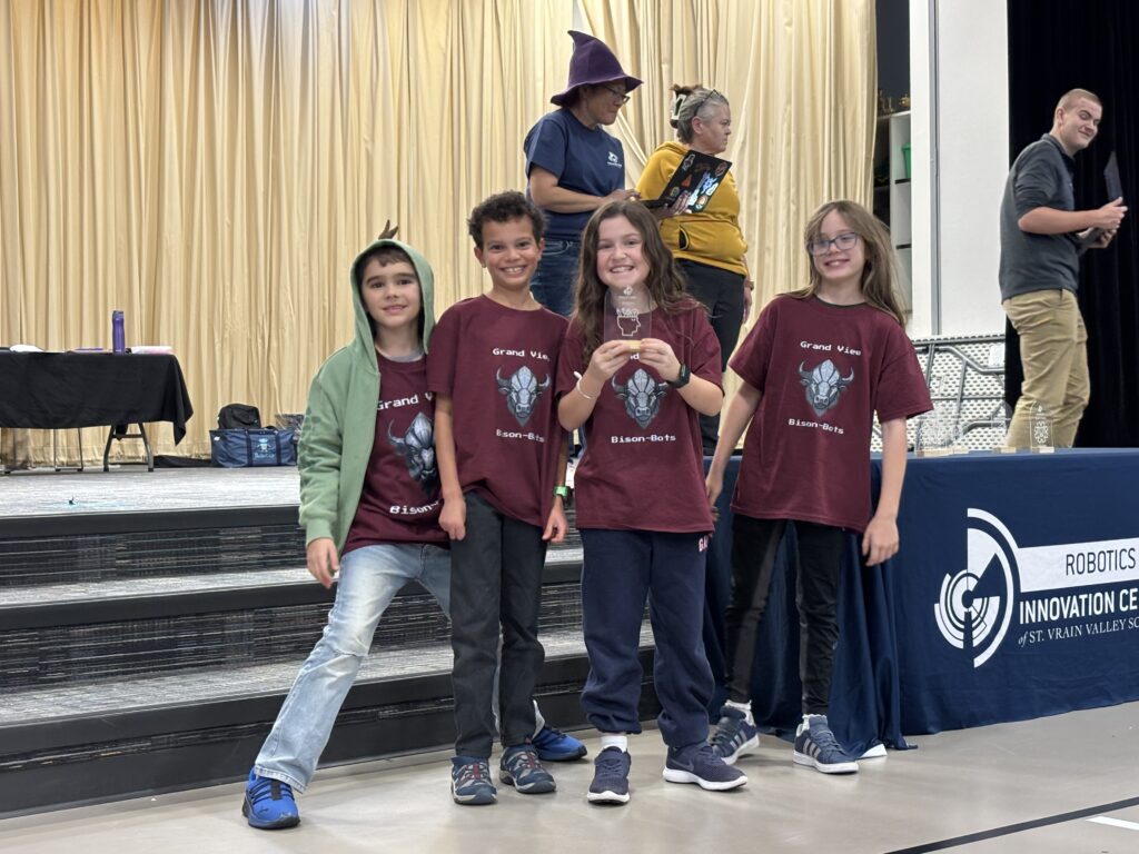 Four children in matching maroon t-shirts pose on stage at an event, smiling and holding a trophy.