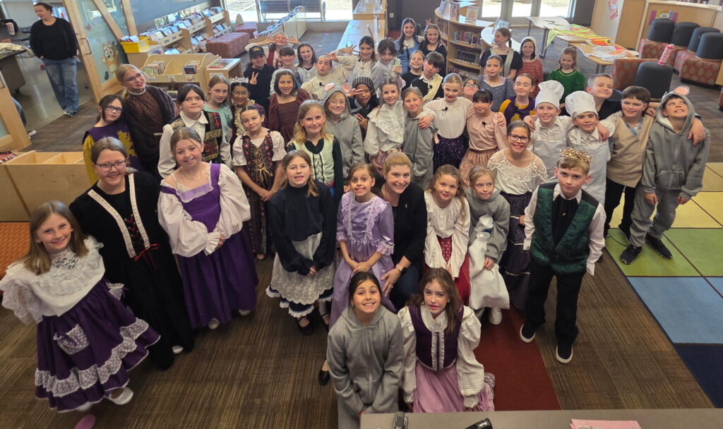 A large group of children in costumes poses together in a library, smiling and enjoying a themed event.