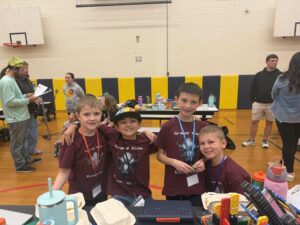 Four boys in matching maroon shirts pose together in a gym, with tables of supplies and people in the background.