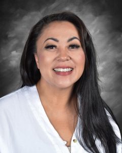 Smiling woman with long dark hair, wearing a white blouse, against a gray backdrop.
