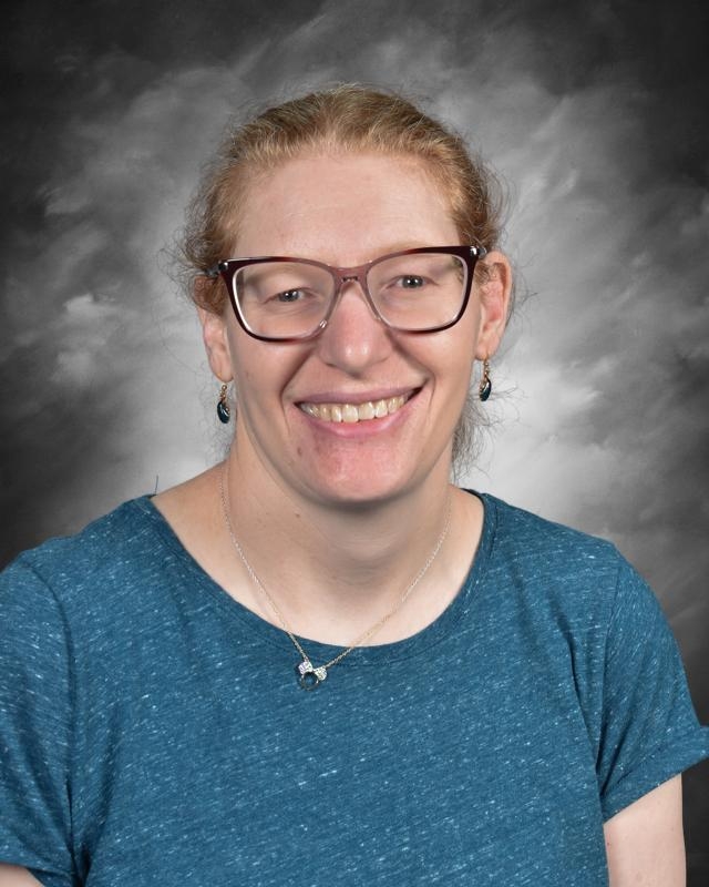 Persona sonriente con gafas, vestida con una camisa azul, sobre un fondo gris.